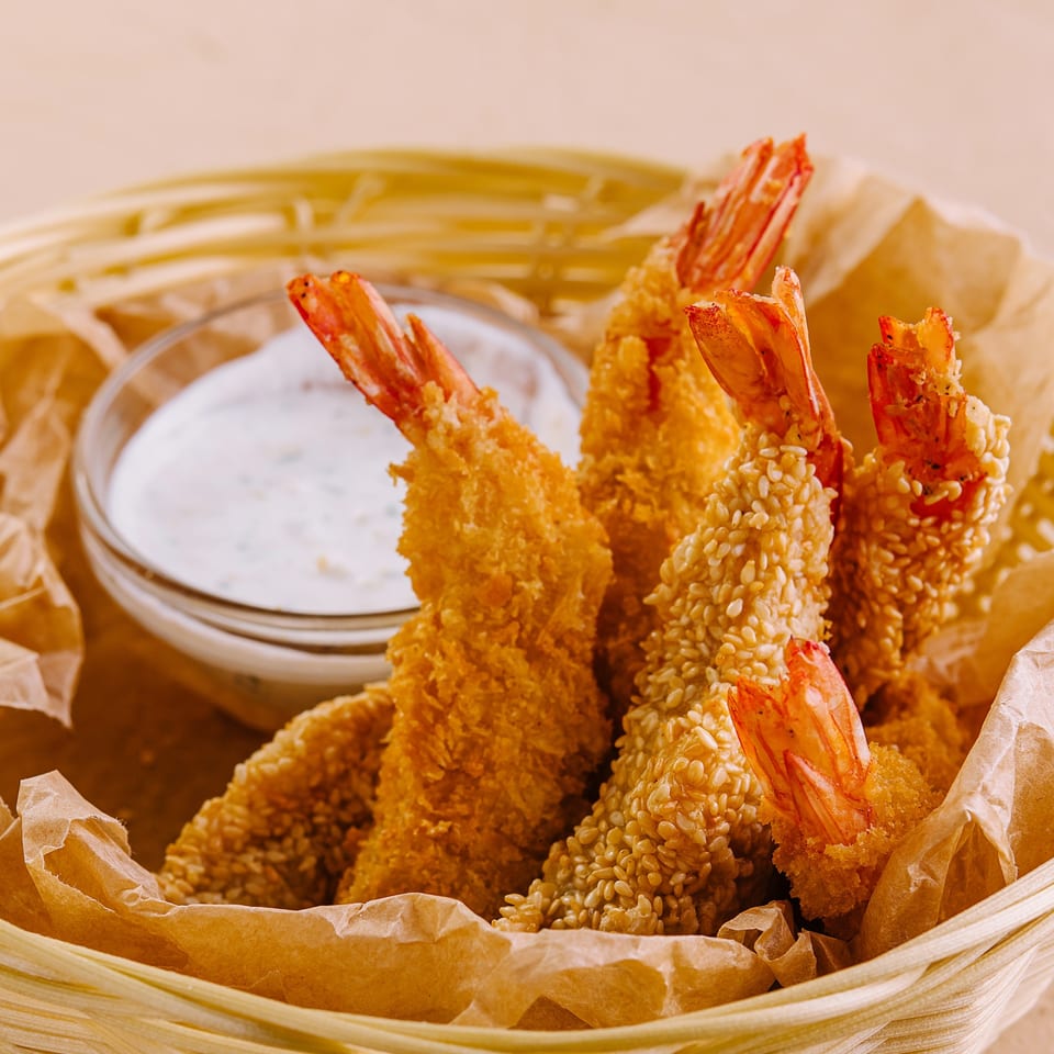 Crispy fried shrimp, some with sesame seeds, and a side of white dipping sauce in a basket.