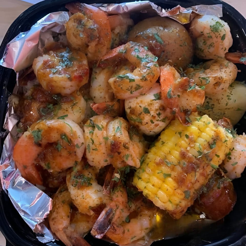 Shrimp, corn on the cob, and potatoes seasoned with herbs in a black bowl lined with foil.