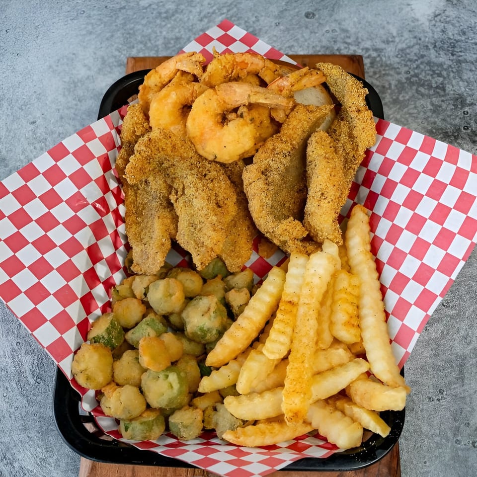 A black basket filled with fried shrimp, fish, okra, and crinkle-cut fries on red and white checkered paper.
